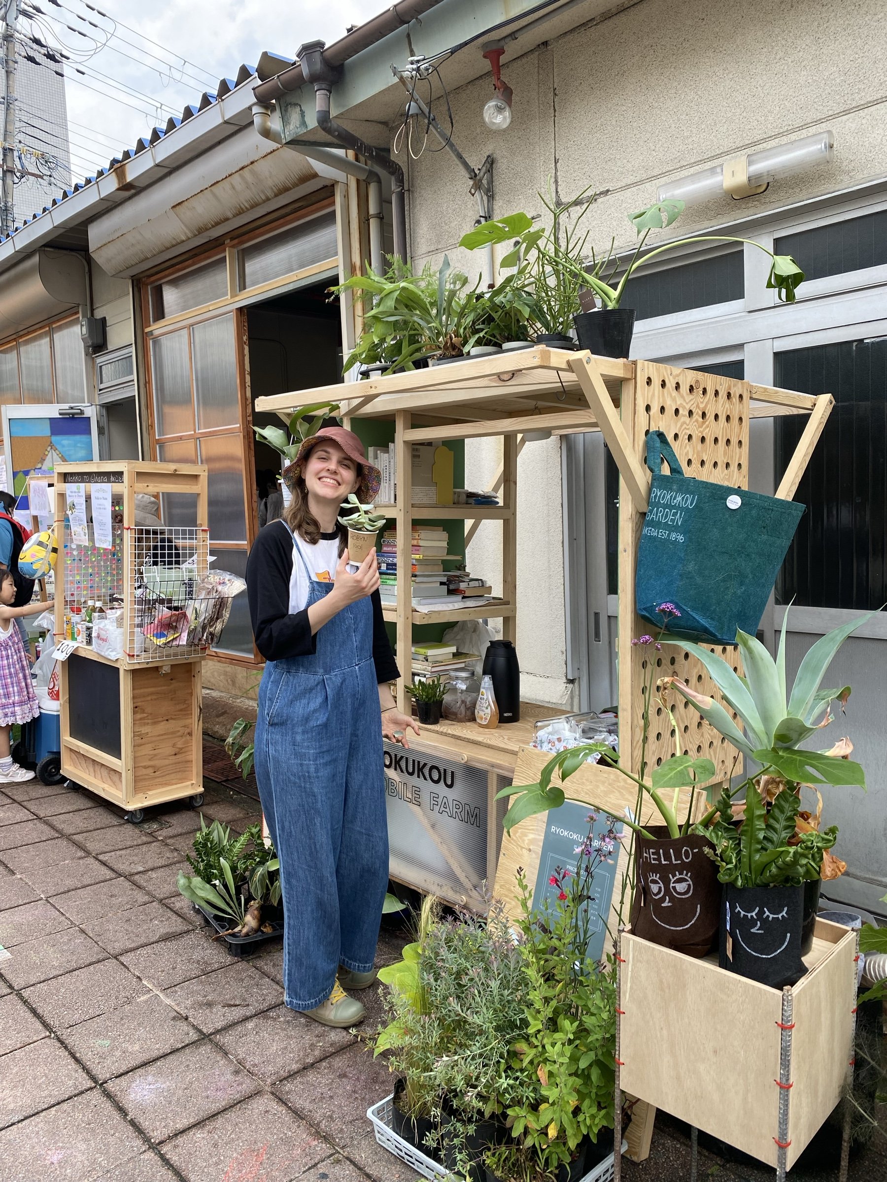 Robyn holding a small potted plant beside a wooden stall of plants and books.