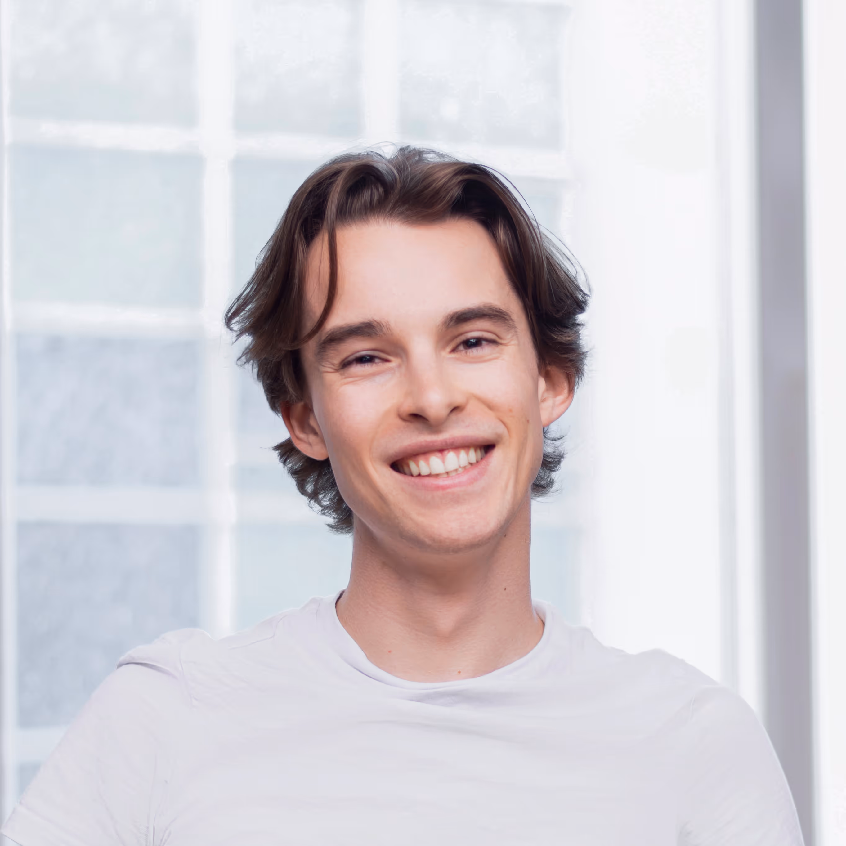 Young man with medium-length brown hair smiling, wearing a white t-shirt against a window background.