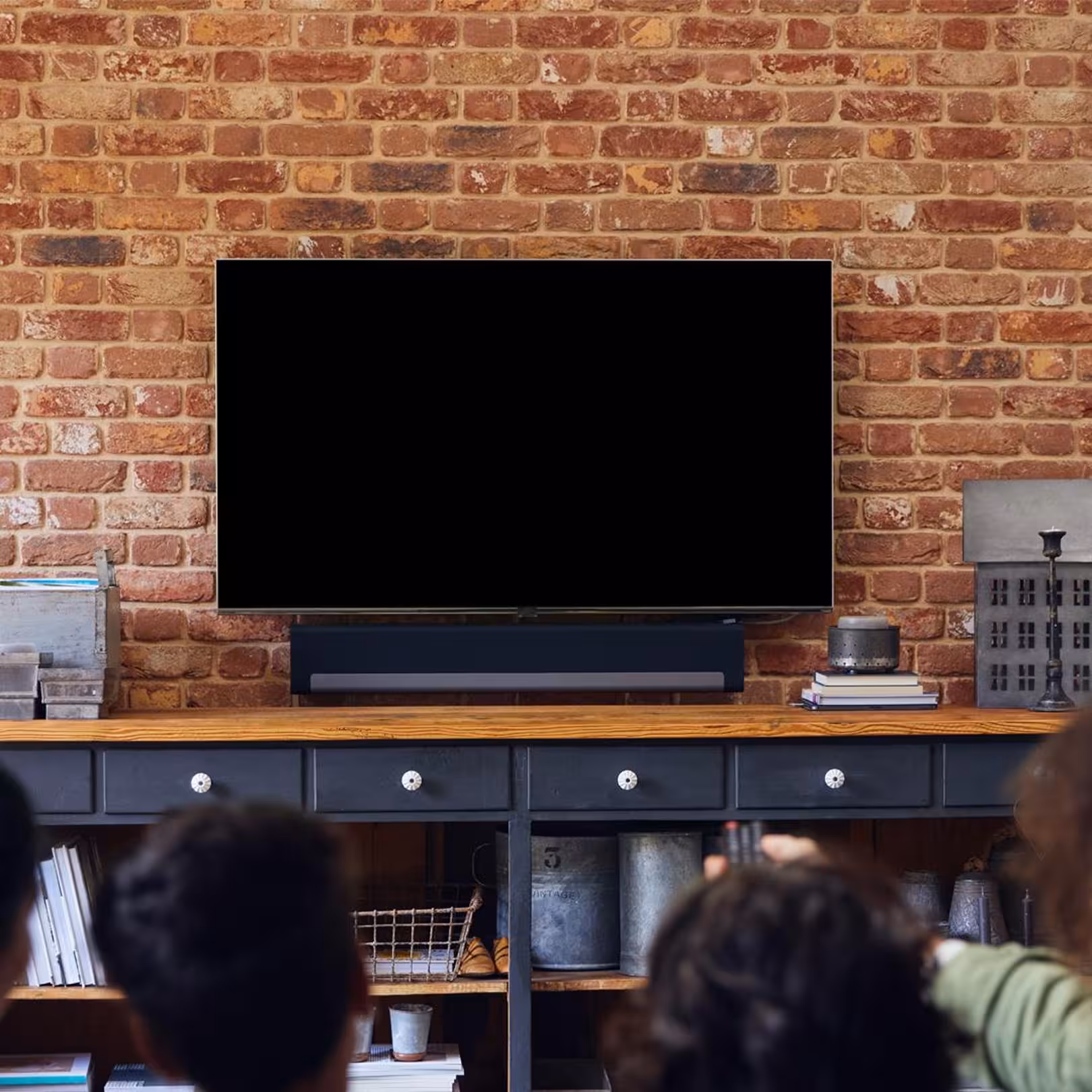 People sitting and watching a blank flat-screen TV mounted on a brick wall above a console table with books and decor.