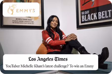 YouTuber Michelle Khare sitting cross-legged on a chair, wearing a red jacket and black boots, with framed Emmy and Black Belt posters behind her.