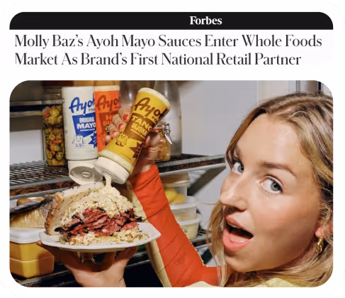 A woman holding a plate with a sandwich piled with pastrami, pouring Ayo! Tangy Dijon mayo sauce on it from a bottle.