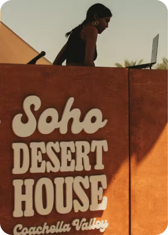 A woman using a laptop behind a wooden podium with 'Soho Desert House Coachella Valley' text on it.