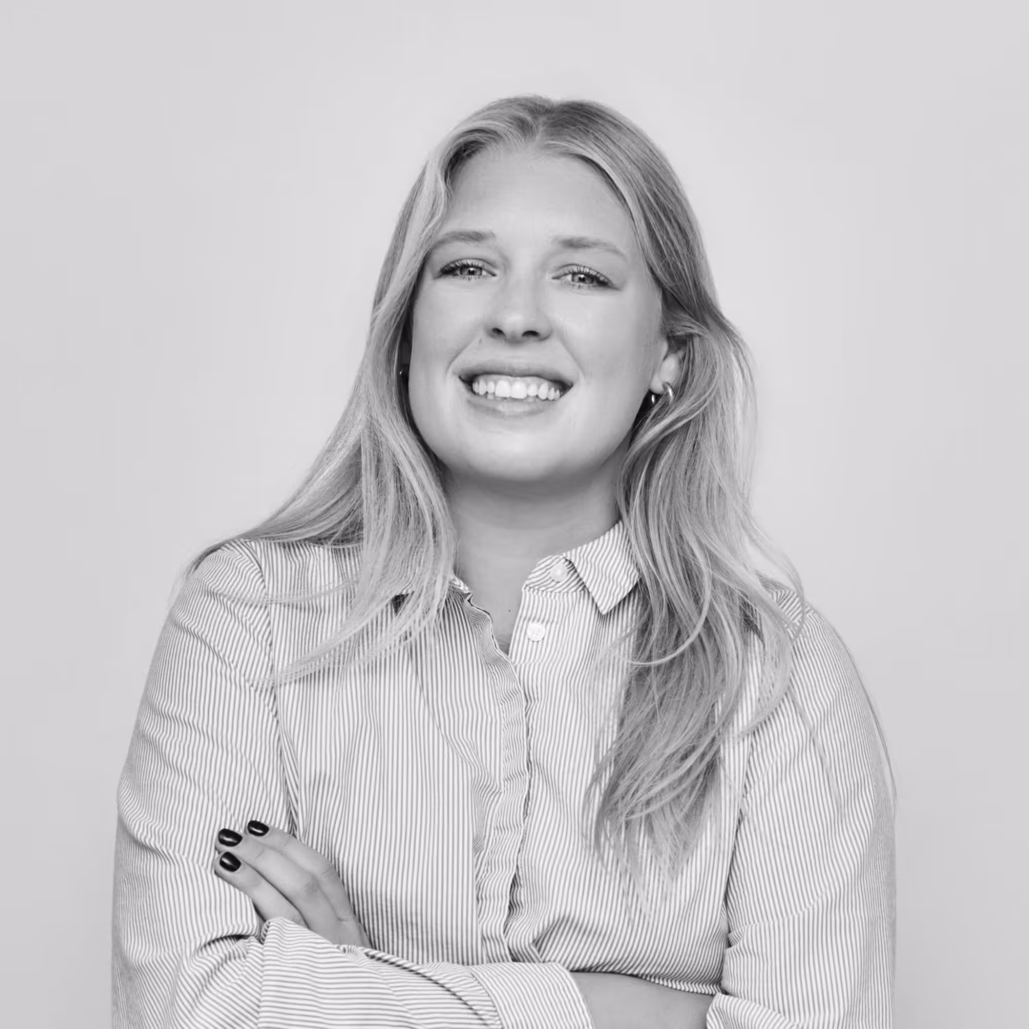 Smiling young woman with long hair wearing a striped shirt and crossed arms against a plain background.