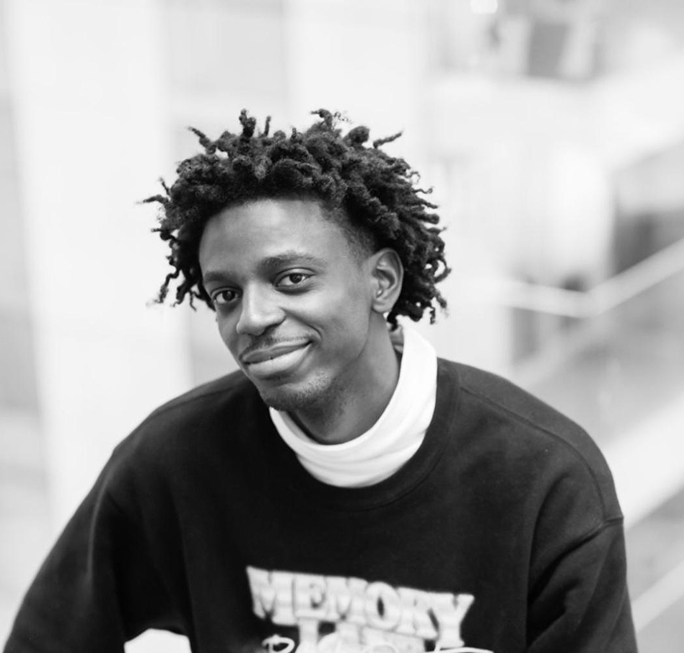 Smiling young man with natural curly hair wearing a black sweatshirt over a white turtleneck indoors.