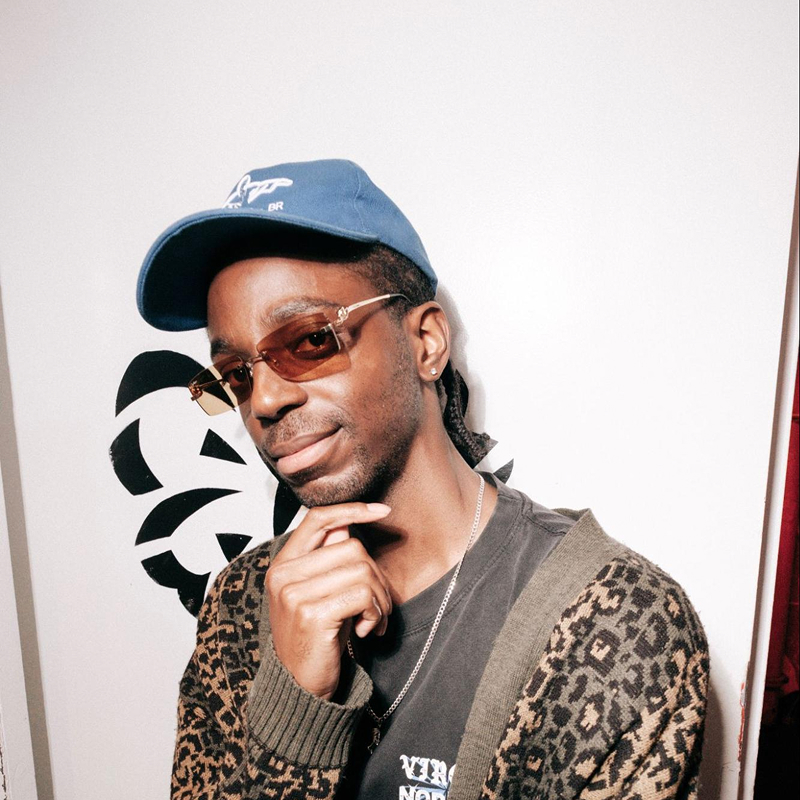 A man wearing a blue cap, brown-tinted sunglasses, leopard print cardigan, and a chain necklace poses with his hand on his chin in front of a white wall with a black geometric design.