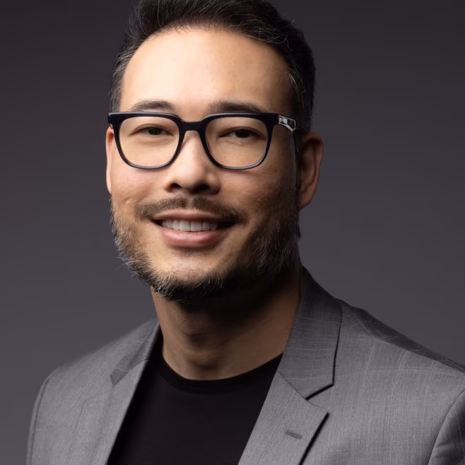 Smiling man with glasses wearing a gray blazer and black shirt on a dark gray background.