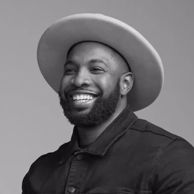 Smiling man with beard wearing a wide-brimmed hat and dark jacket on a plain background.