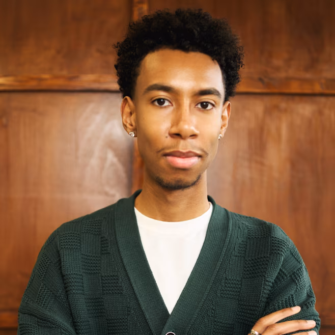 Young man with short curly hair wearing a green sweater over a white shirt, standing with arms crossed.