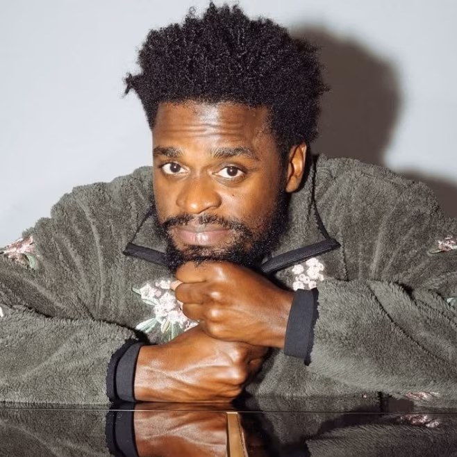 Man with beard and afro hairstyle leaning on polished surface, wearing green floral fleece jacket.