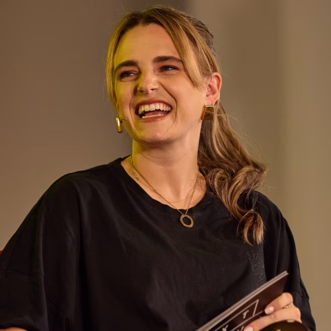 Smiling woman with blonde hair tied back, wearing a black top and holding a booklet.