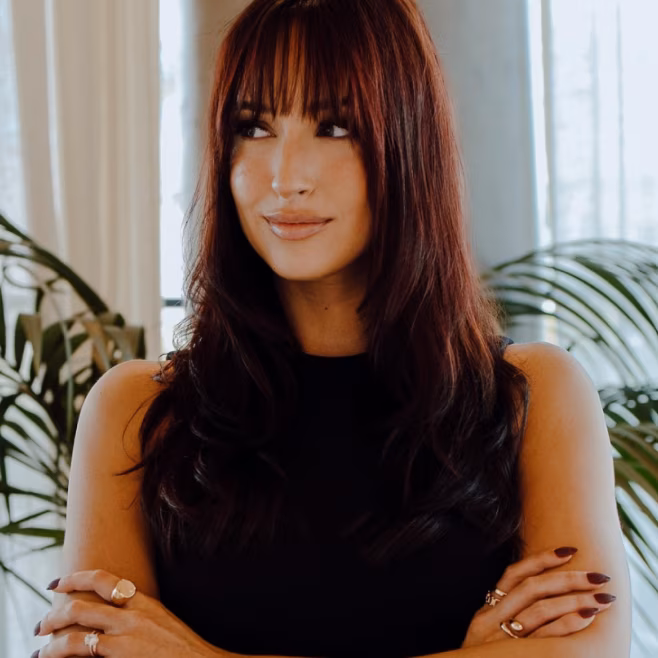 Woman with long dark hair and bangs, arms crossed, looking to the side with a slight smile indoors.