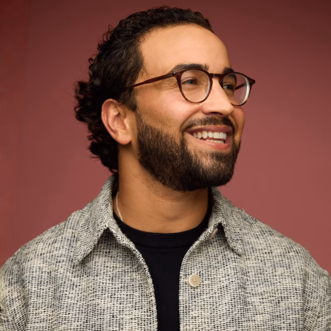 Man with beard and glasses smiling and looking to the right against a muted red background.