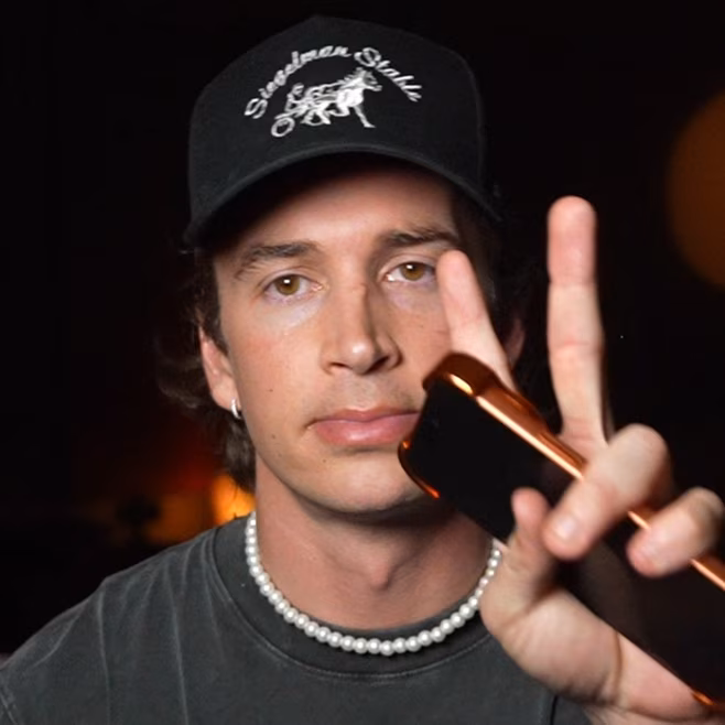 Man wearing a black cap and pearl necklace, holding a phone while making a peace sign gesture.
