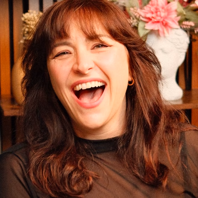 Woman with brown hair laughing with mouth open, wearing a black top, with flowers in the background.