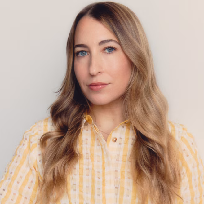 Woman with long wavy blonde hair wearing a yellow patterned shirt against a plain background.