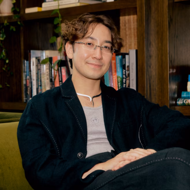 Young man with glasses and curly hair sitting on a green sofa with bookshelves behind him.