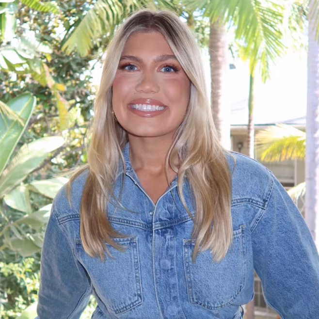 Smiling woman with long blonde hair wearing a blue denim jacket outdoors with green foliage behind.