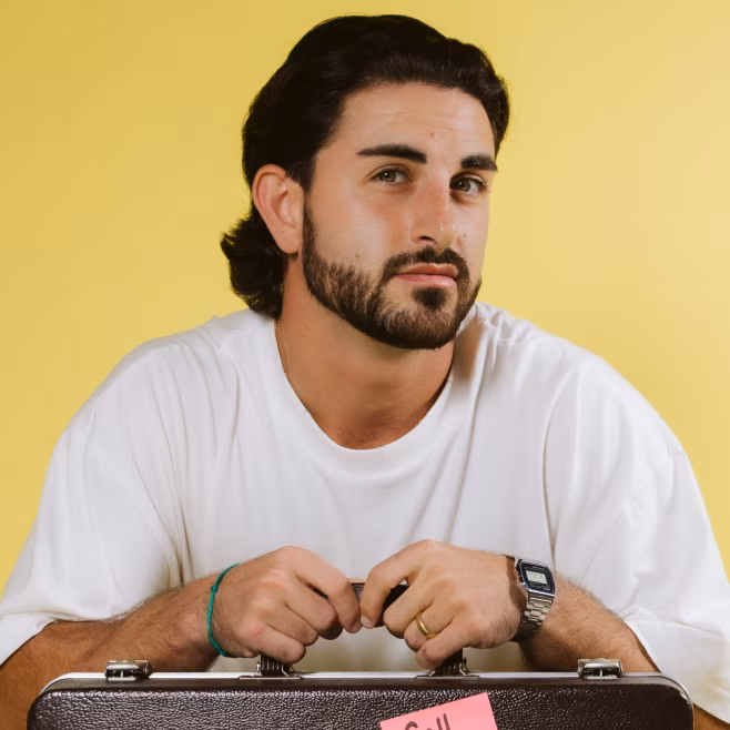 Man with dark hair and beard wearing a white shirt, leaning on a briefcase against a yellow background.