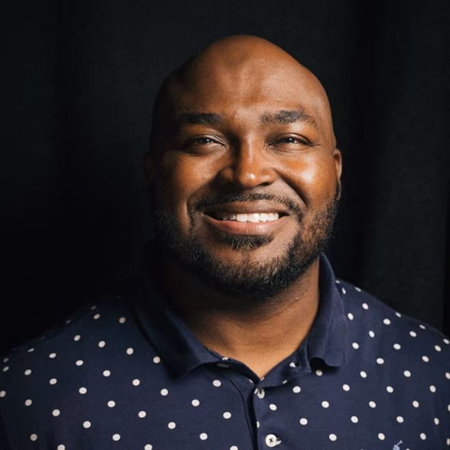 Smiling bald man with a beard wearing a navy blue polka dot shirt against a dark background.