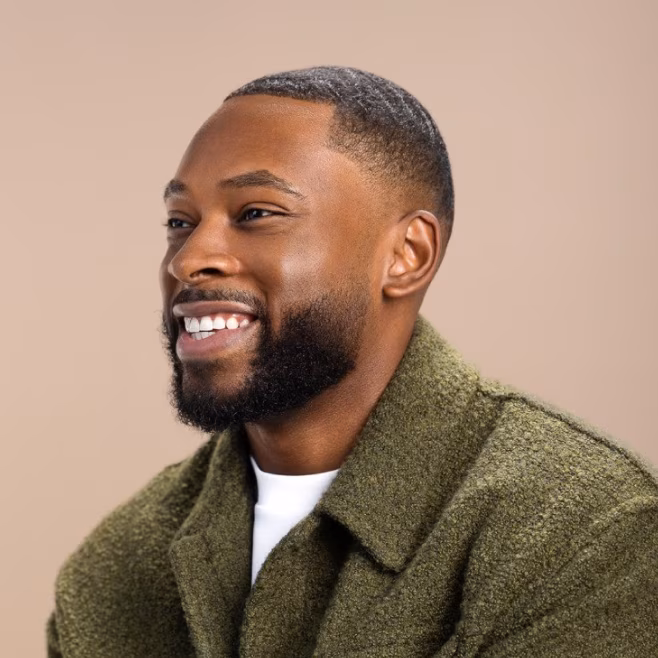 Smiling man with beard and short hair wearing a green coat against a beige background.