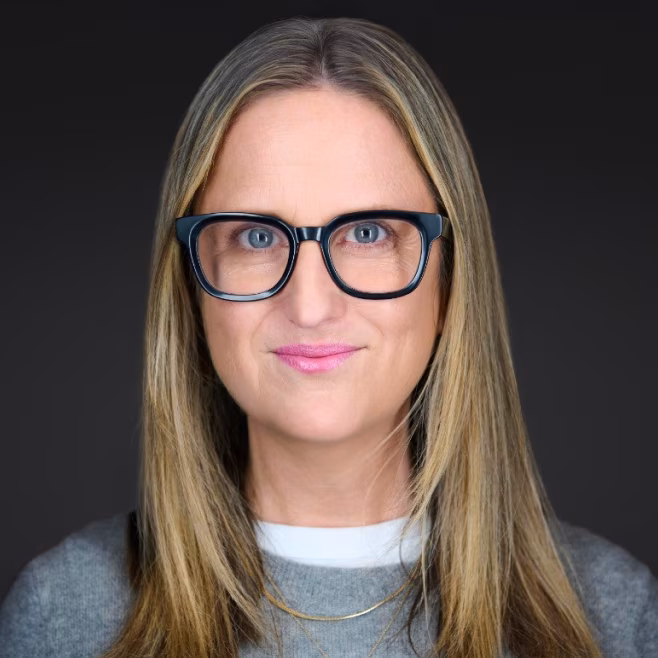 Woman with long blonde hair and black framed glasses wearing a gray sweater and white shirt.