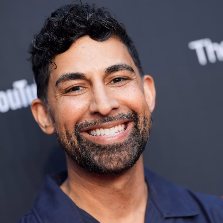 Portrait of a smiling man with curly black hair and a beard against a dark background.