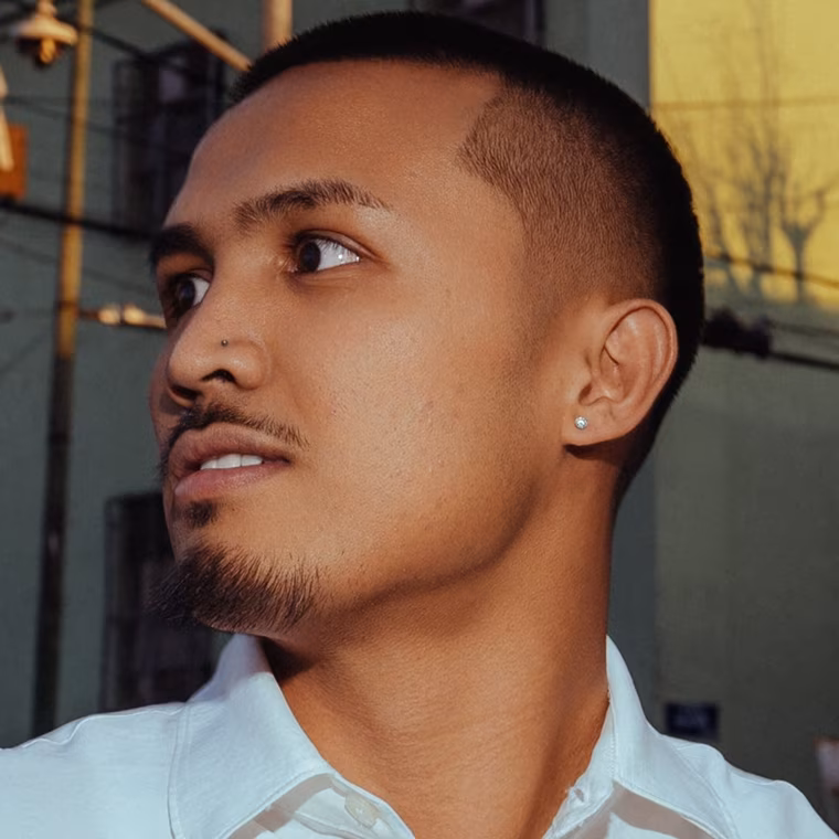 Close-up of a man with a goatee, nose and ear piercings, wearing a white shirt, looking left.