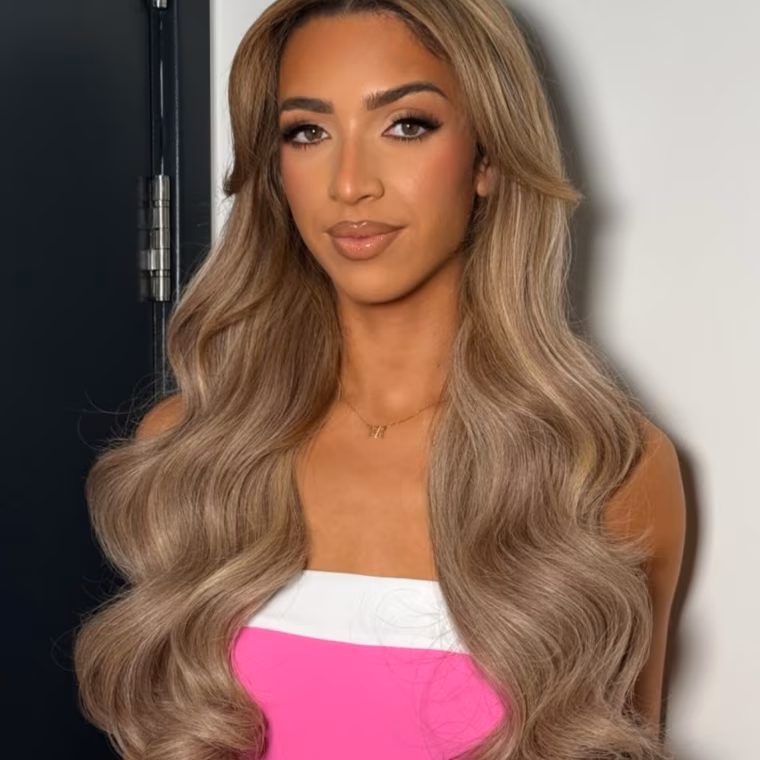 Woman with long wavy blonde hair wearing a pink and white top against a plain background.