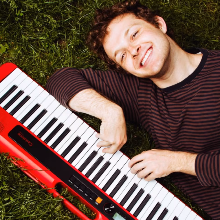 Smiling man in striped shirt lying on grass playing a red and black keyboard.
