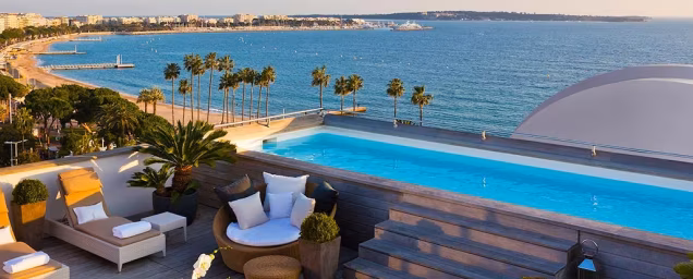 Rooftop pool with seating overlooking a beach, palm trees, and ocean under a clear sky.