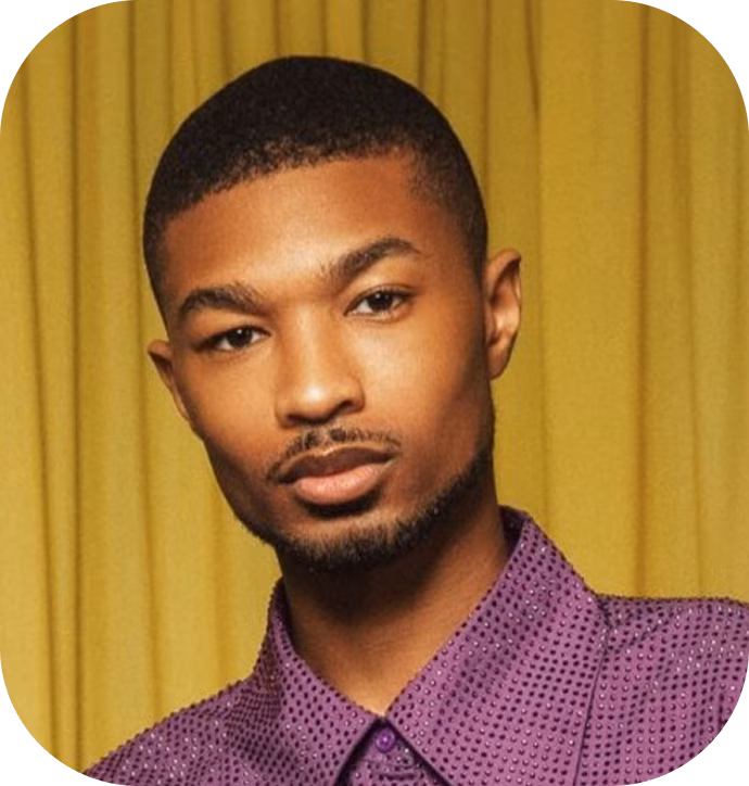 Portrait of a young man with short hair and a trimmed beard wearing a purple shirt with small dots, set against a yellow curtain background.