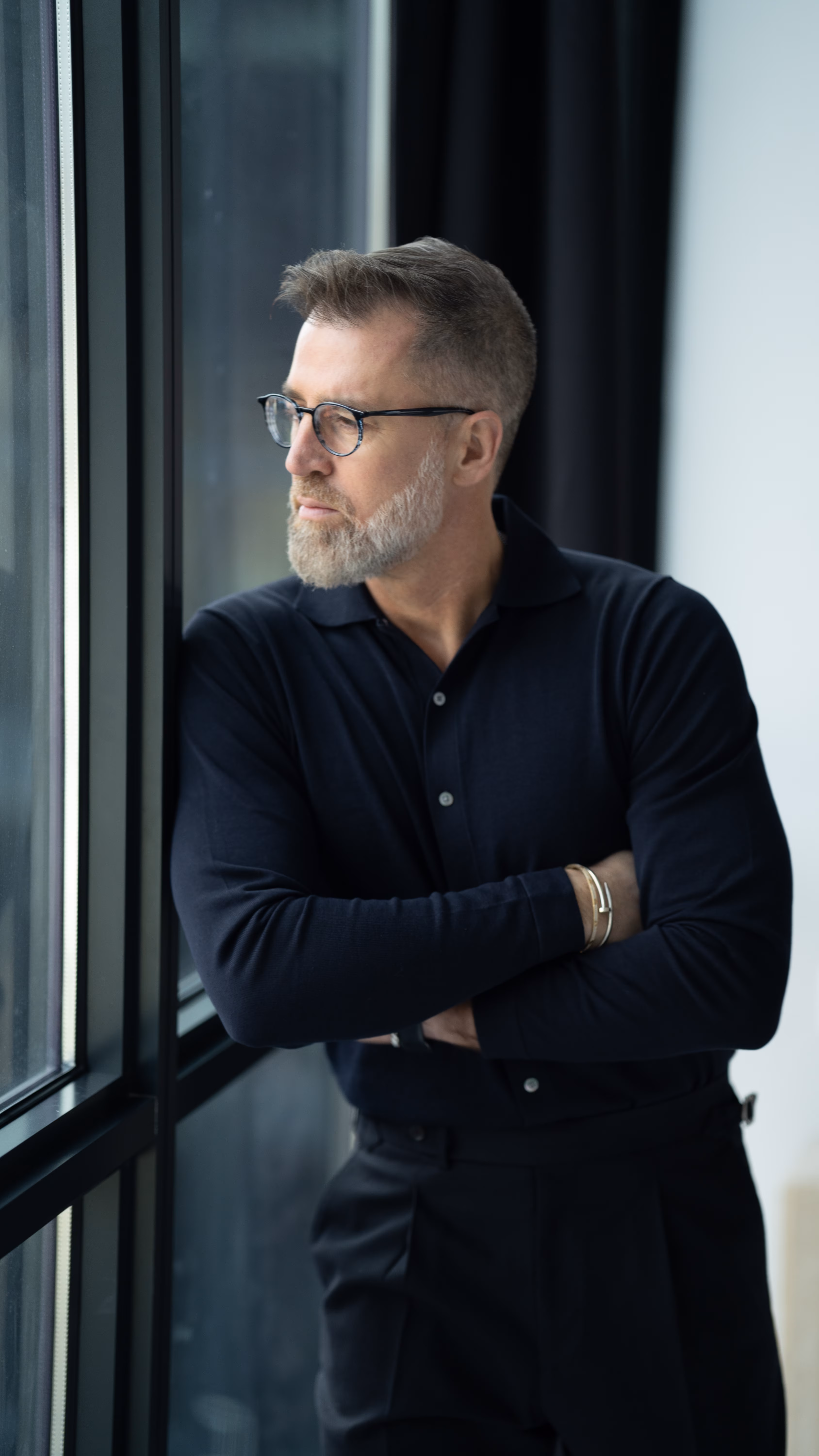 A middle-aged man with gray hair and beard wearing glasses and a black button-up shirt stands with arms crossed, gazing out a large window.
