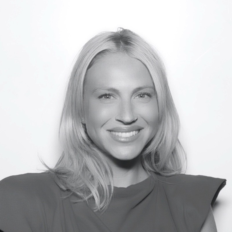 Portrait of a smiling woman with shoulder-length blonde hair wearing a dark top with broad shoulder details.