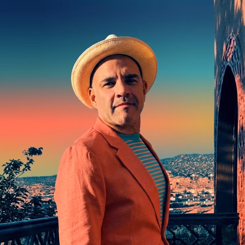 Man wearing a beige straw hat, orange blazer, and teal striped shirt standing outdoors with a scenic city and hills in the background during sunset.