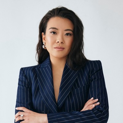 Confident woman with long black hair wearing a navy blue pinstripe blazer, standing with arms crossed against a plain light background.