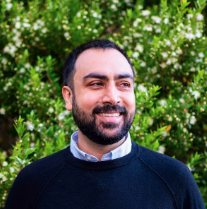 Man with short dark hair, beard, and mustache wearing a black sweater over a collared shirt, smiling and looking slightly to the side, standing outdoors with green leafy bushes in the background.