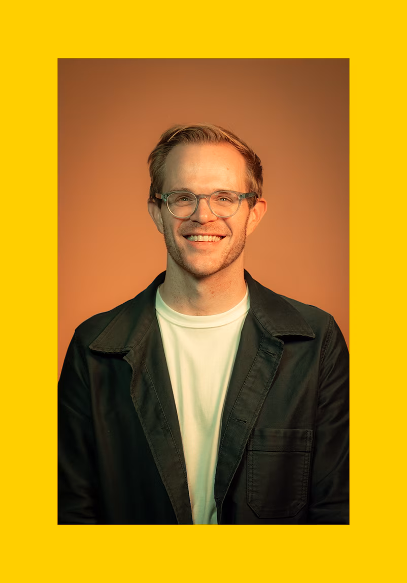Smiling young man with short light brown hair wearing glasses, a white t-shirt, and a dark jacket, posed against a warm brown background with a bright yellow border.