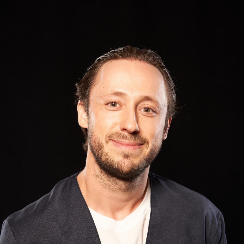 Man with light skin tone, short brown hair, and beard, wearing a dark blazer over a white shirt, smiling gently against a black background.