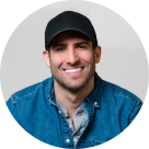 Smiling man wearing a black baseball cap and a blue denim jacket over a gray hoodie.