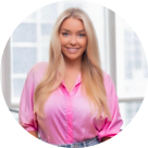 Smiling woman with long blonde hair wearing a bright pink satin blouse, posed in front of a bright window.