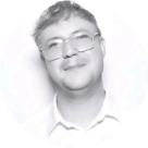 Black and white photo of a young person smiling, wearing glasses and a collared shirt, with short curly hair.