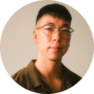 Headshot of a young person with short black hair and glasses, wearing a brown shirt.