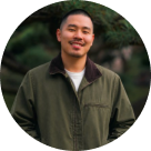 Smiling man with short hair and mustache wearing an olive green jacket over a white shirt, standing outdoors with blurred natural.