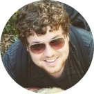 Smiling person with curly hair wearing sunglasses and a dark shirt.
