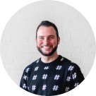 Smiling bearded man with short dark hair wearing a black shirt patterned with white hashtags, standing against a white brick wall.