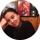 A smiling woman with dark hair pulled back, resting her head on her right hand, sitting in a room with wooden walls and framed pictures behind her.
