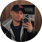 Young man taking a mirror selfie with a smartphone, wearing a black cap with a red logo, black shirt, silver chain, and a jacket with a gray hood, standing in a kitchen.
