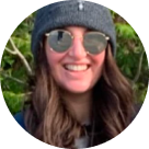 Smiling person wearing round reflective sunglasses, a gray beanie hat with a small logo, and a dark jacket, with greenery.