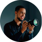 Smiling man with short curly hair and a beard holding a DSLR camera, looking slightly to the side.