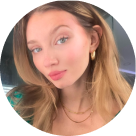 Close-up portrait of a young woman with light skin and long blonde hair, wearing gold hoop earrings and layered gold necklaces, looking at the camera with a slight smile.